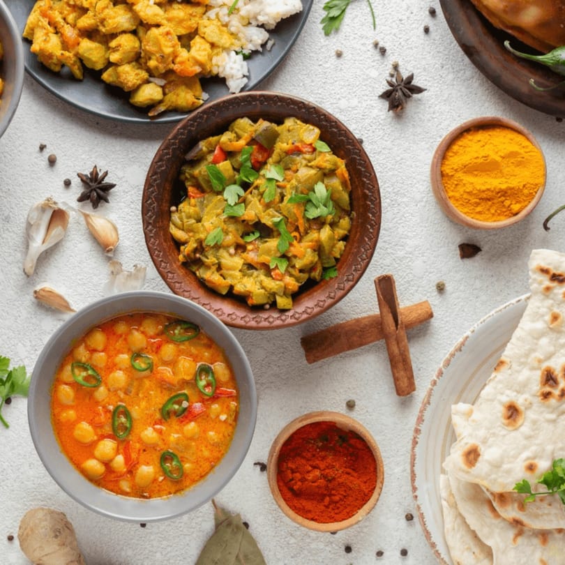 Overhead view of various Indian dishes including chickpea curry, chicken, vegetables, naan, and spices.
