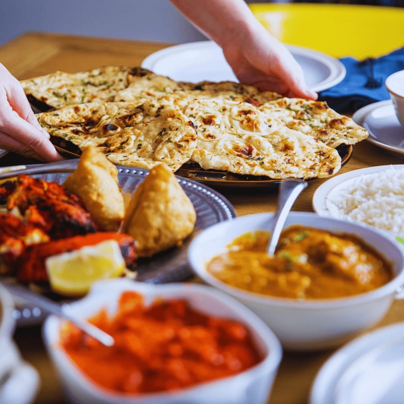 Hands serving naan bread alongside various Indian dishes like samosas, tandoori chicken, and curries.
